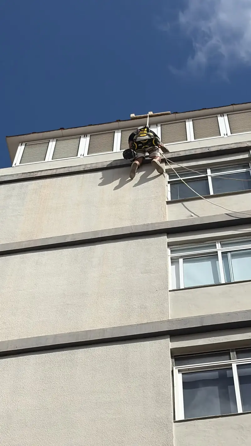 Trabajador colgado en lo alto de una fachada gris con el cielo azul